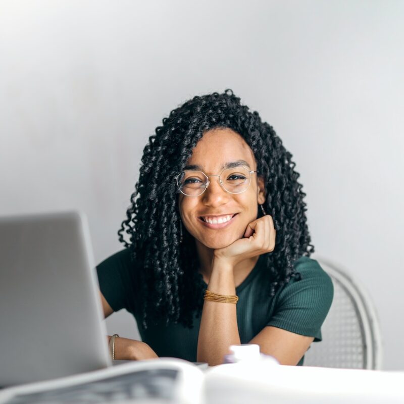Ragazza mora con capelli ricci e occhiali da vista che sorride con mano sotto il mento. Alle spalle ha una parete grigia e di fronte un pc grigio adagiato su un tavolo con un libro bianco aperto. Ha completato il metodo pomodoro e si sta rilassando.