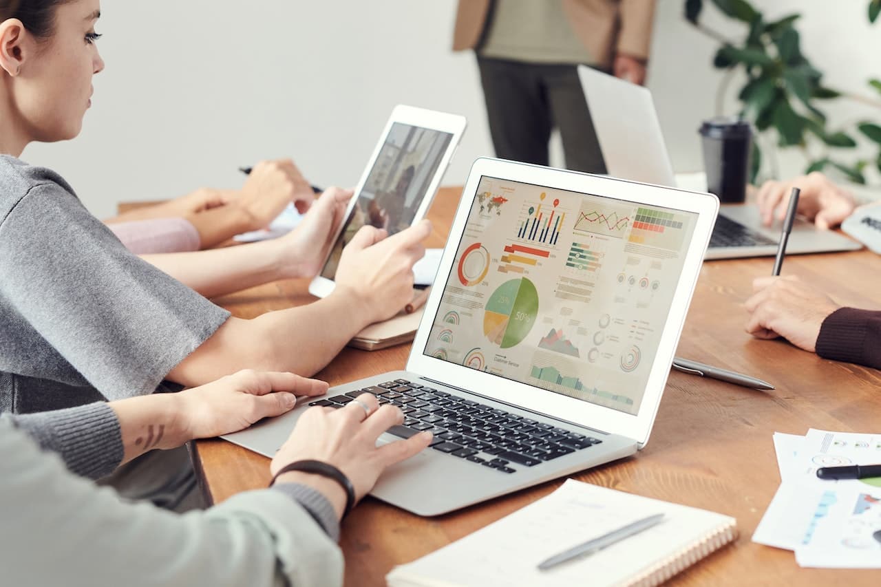 Tavolo in legno a cui sono seduti degli analisti con computer, tablet, penne e quaderni. Sulla sinistra, in primo piano ci sono delle mani che usano un pc, che ha sul desktop molti grafici e indici di economia. Di fianco c'è una ragazza di profilo che tiene in mano un tablet su cui sta visualizzando altri dati.