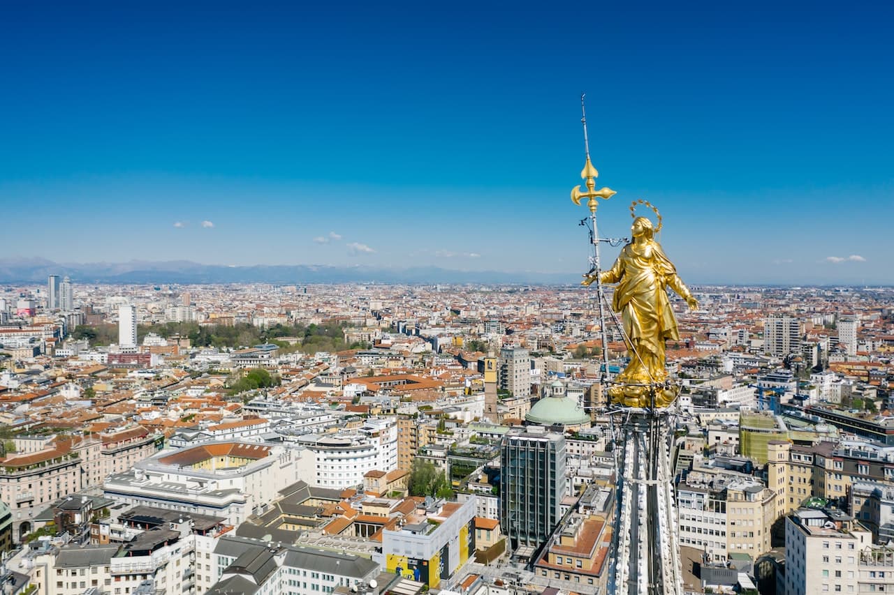 In primo piano c'è la Madonnina del Duomo di Milano, città con molte università per studiare economia. Sullo sfondo c'è il cielo senza nuvole e di gradiente azzurro verso l'altro. Tra la Madonnina e il cielo sono visibili molti edifici del centro della città.