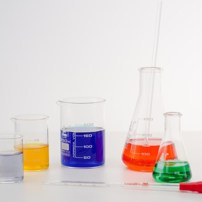 Sfondo bianco con ampolle e becker di laboratorio in primo piano. Ognuno di questi contiene un liquido diverso con specifiche proprietà periodiche.