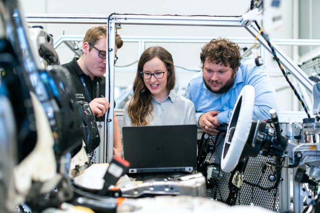 Tre studenti delle migliori università di ingegneria che lavorano su un progetto, mentre sono di fronte un pc e circondati da elettronica e pezzi metallici. Sono due ragazzi e una ragazza (al centro).