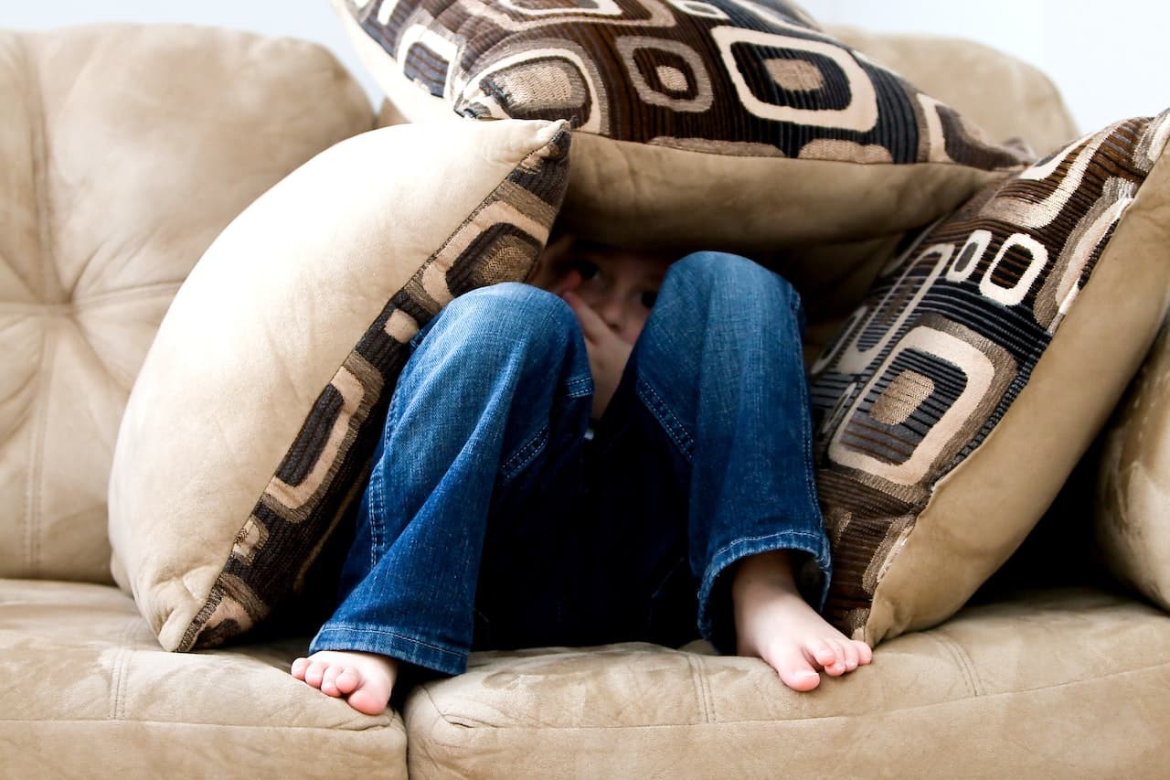 Bambino che si nasconde sotto i cuscini di un divano di colore beige. In primo pianto spuntano le gambe con i pantaloni di jeans. Il bambino si nasconde perché deve affrontar le sue paure e contrastare l'ansia da esame.