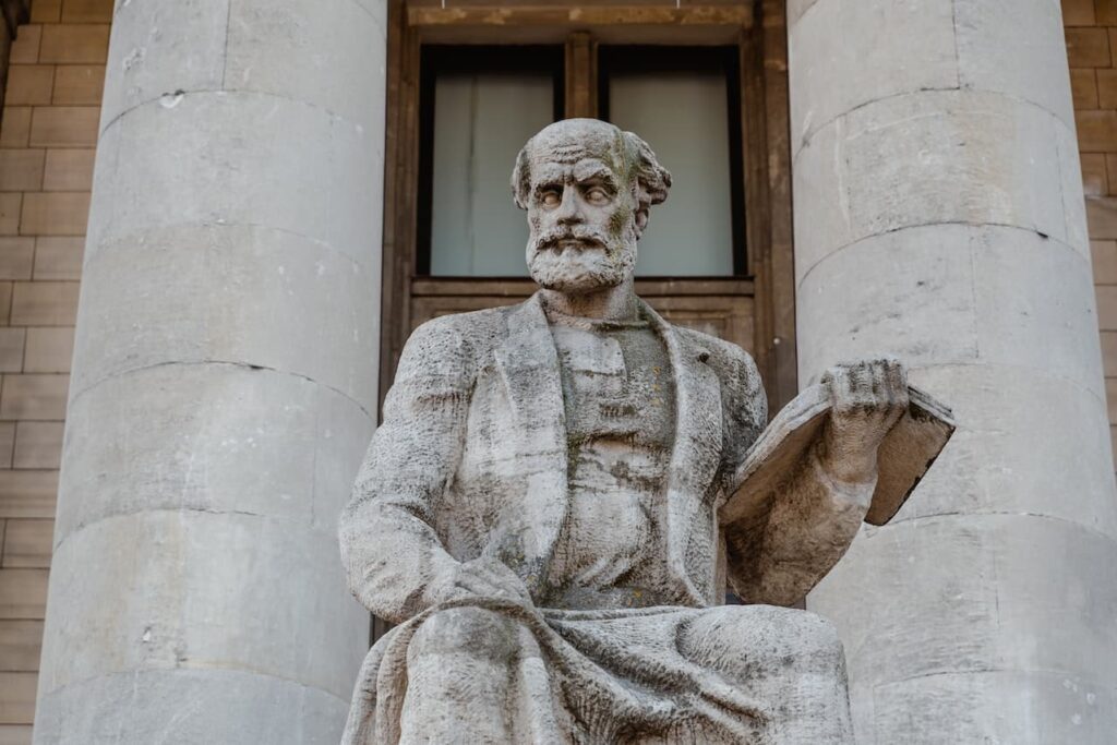 Statua i n pietra di un uomo barbuto sulla 60na. Sullo sfondo ci sono due colonne alle spalle dell'uomo.