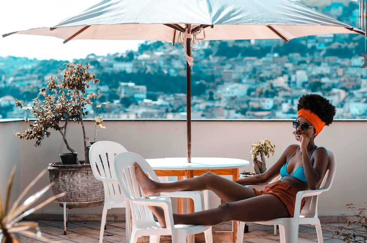 Donna in costume da bagno seduta su una sedia bianca sotto un ombrellone bianco in un patio. Ha le gambe distese su un altra sedia bianca e sullo sfondo ci sono delle case sfocate. La donna si sta rilassando.