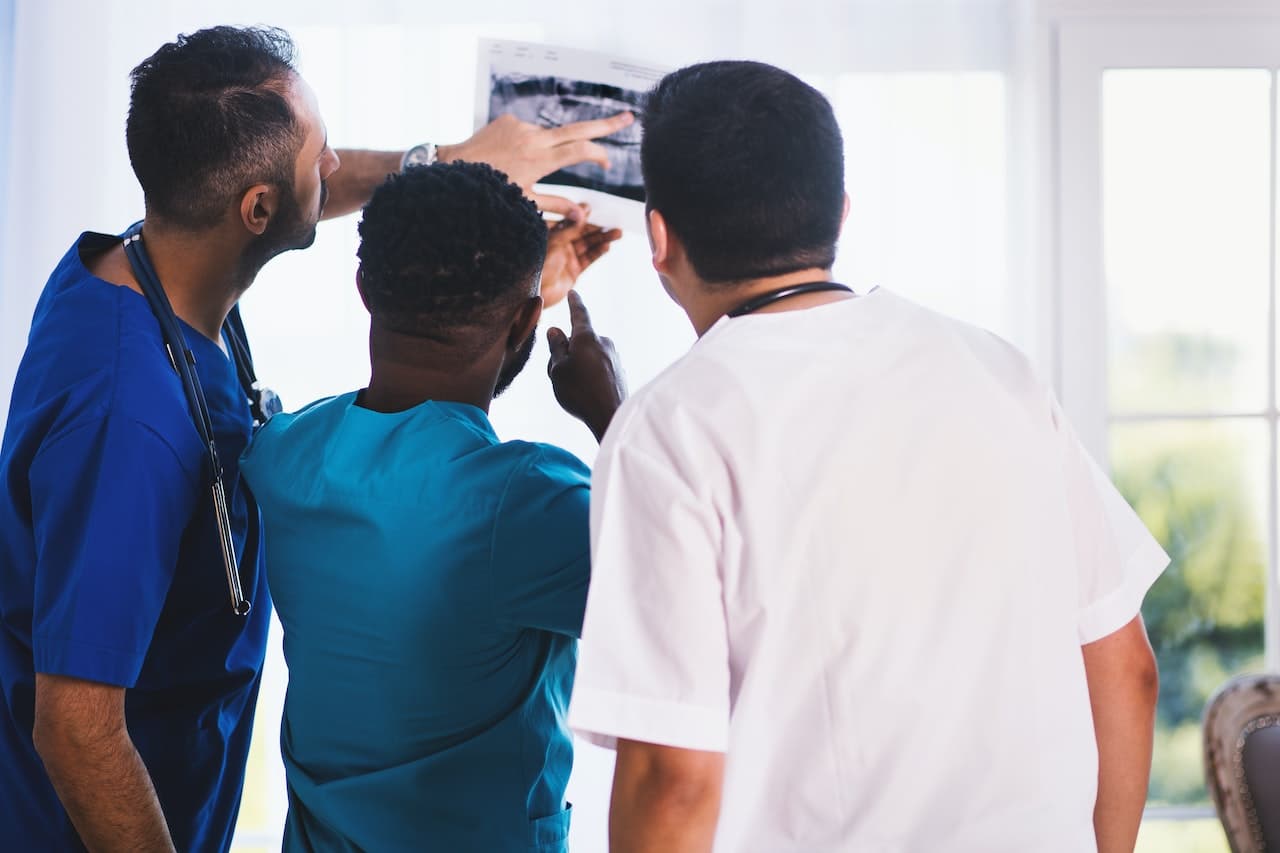 Chi è il fisiatra e cosa fa oltre a studiare per la specializzazione? Il fisiatra deve gestire (in modo conservativo) disturbi relativi sia all'apparato locomotore che ai nervi periferici. Nell'immagine ci sono 3 medici con camici di colore blu, verde e bianco (da sinistra a destra) che guardano una lastra di un paziente.