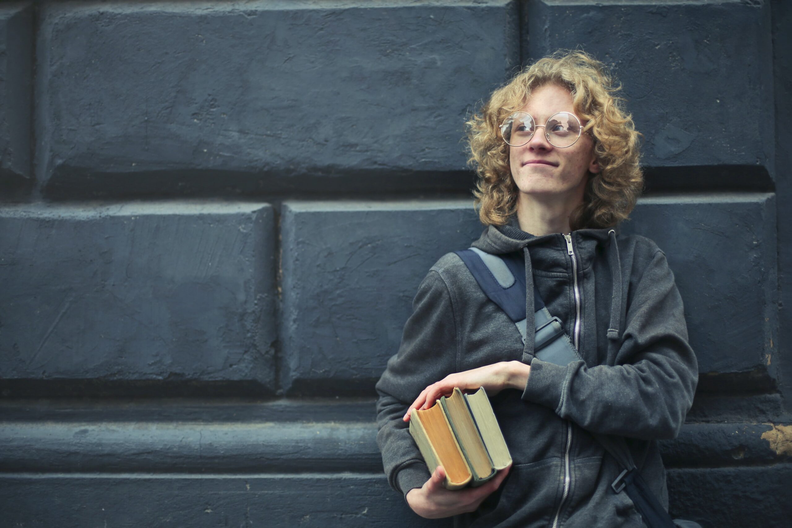 Studente con tre libri sotto il braccio e poggiato a un muro dai mattoni neri.