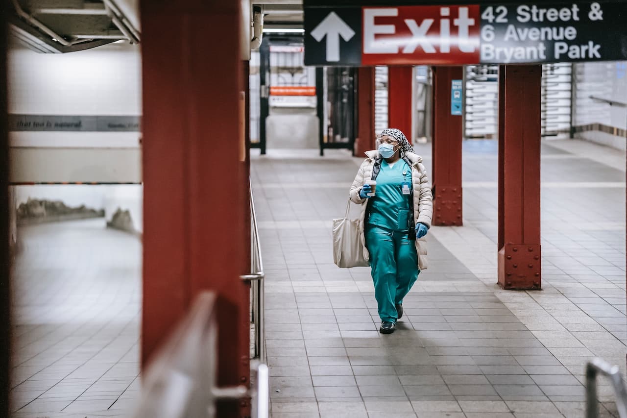 Infermiere che cammina all'interno di una fermata della metropolitana.Indossa il completo verde, i guanti, la cuffia per i capelli, la mascherina e ha un cappotto e una borsa bianchi. La figura è in mezzo a colonne rosse.