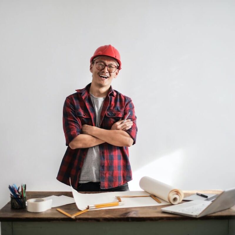 Le formule di fisica per il TOLC permettono un ripasso immediato e veloce. Nell'immagine c'è un ingegnere con le braccia conserte e il volto sorridente. Indossa un casco di sicurezza di colore rosso, una camicia a quadri rossa e nera. Di fronte ha una scrivania con fogli e strumenti per utilizzare le formule della fisica.