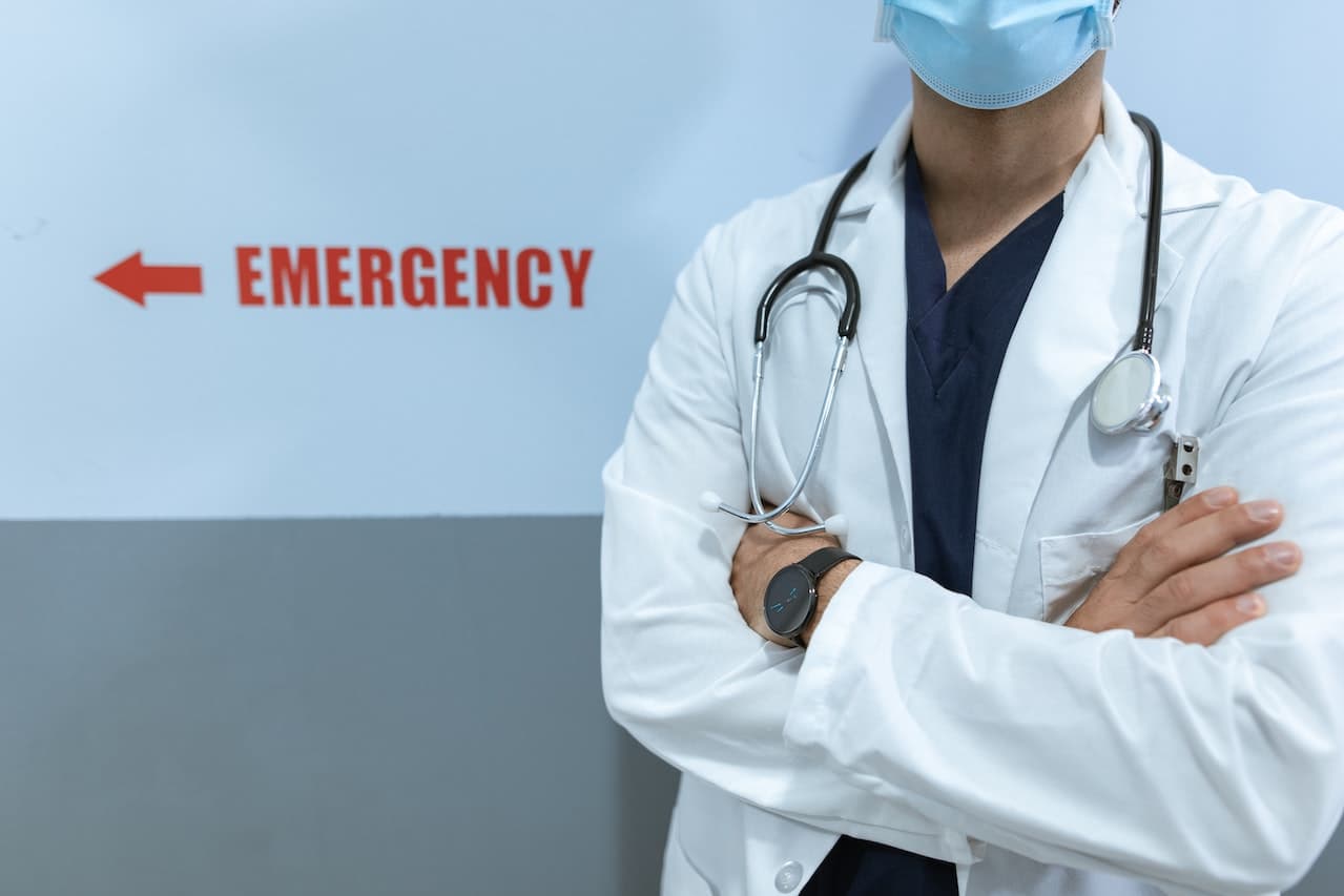 Neurochirurgo con le braccia conserte. Sullo sfondo una scritta emergency in rosso. Il medico indossa un camice bianco, al collo ha un caleidoscopio e indossa una mascherina celecte.