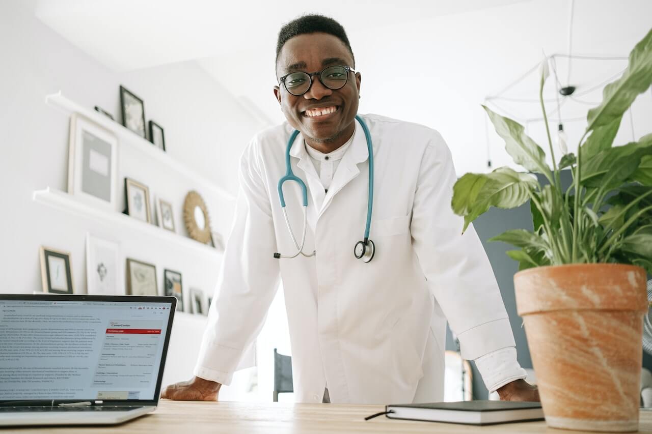 Medico nel suo studio. Indossa il camice bianco e ha al collo uno steitoscopio color indaco.