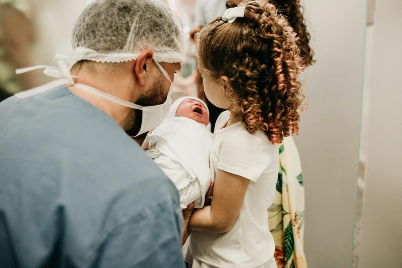Bambino appena nato tenuto in braccio da una bambina e da un infermiere, entrambi girati di spalle.
