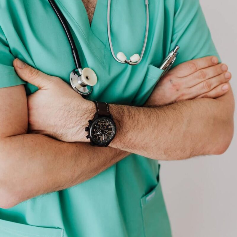 Infermiere con indosso uno scrub verde e lo stetoscopio intorno al collo. Tiene le braccia conserte e indossa un orologio nero sul polso.