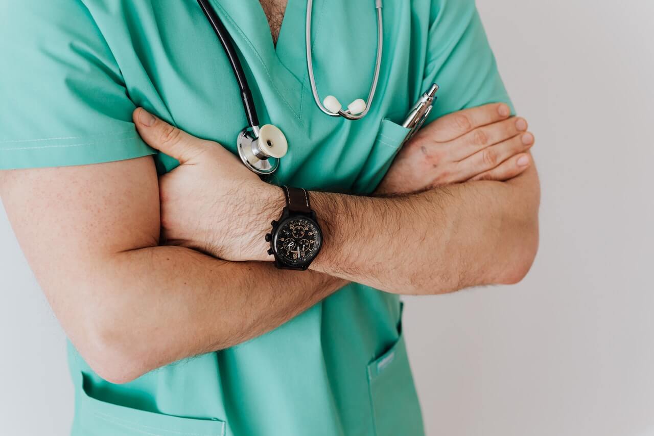 Medico con camice verde e stetoscopio intorno al collo. Ha le braccia conserte e un orologio nero al polso sinistro.
