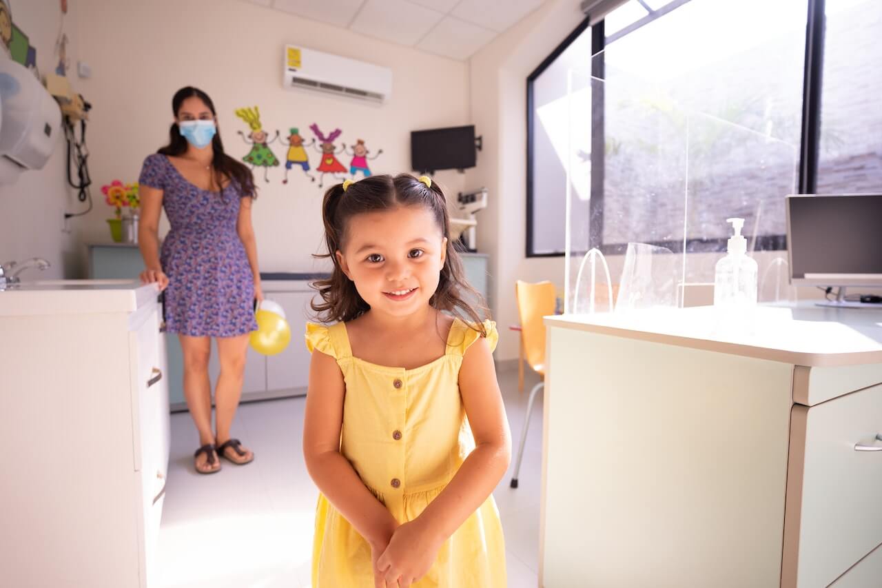 Bambina nello studio del pediatra. Sullo sfondo si vede lo studio medico e la madre della bambina.