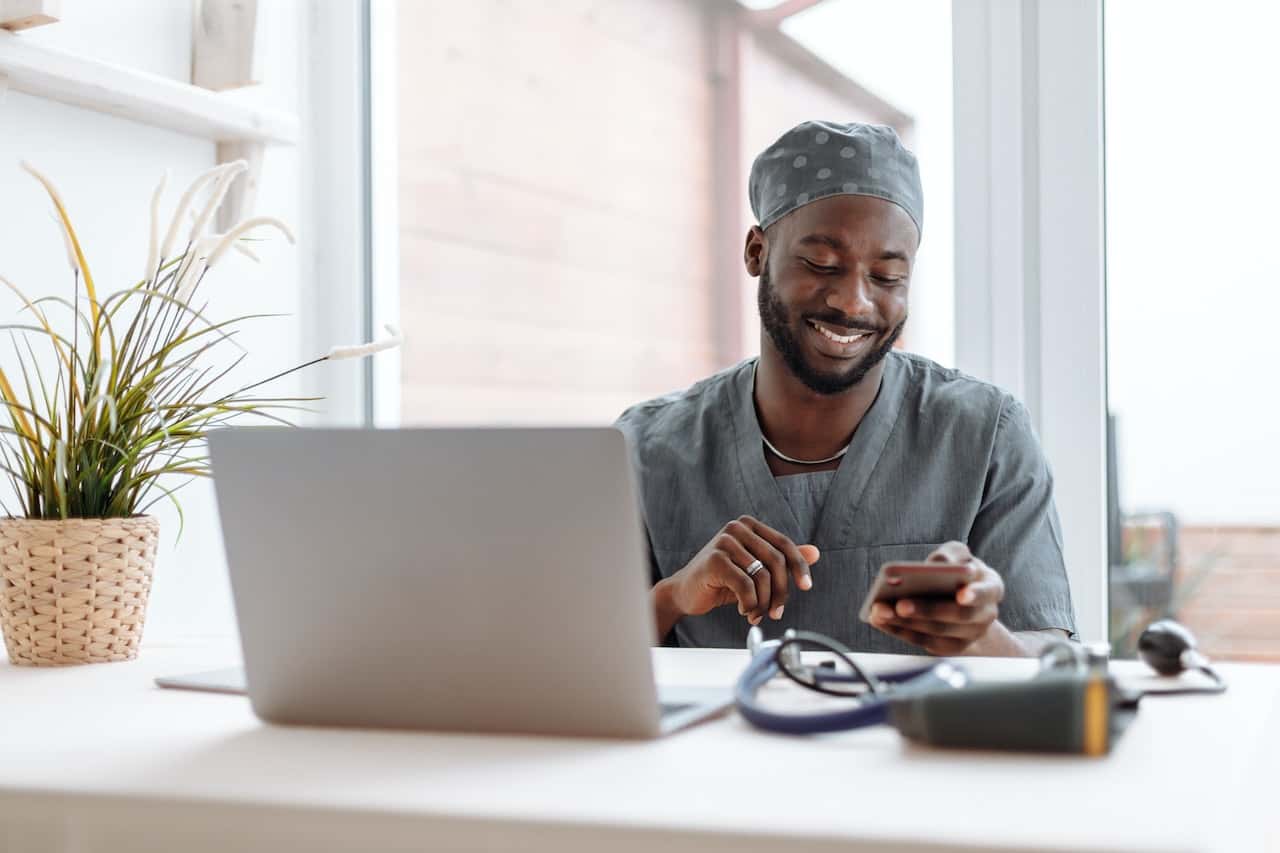 Psichiatra con divisa grigia davanti al pc sulla sua scrivania. Sta guardando il telefono e sorride. Di fianco al computer c'è un caleidoscopio.