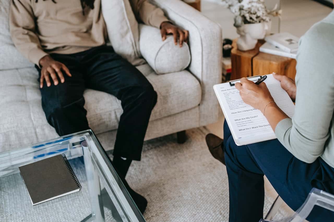 Due persone sedute una di fronte all'altra. Una indossa pantaloni neri e maglia beige ed è seduta sul divano. L'altra indossa pantaloni blu e una maglia grigia, è seduta su una sedia di plastica trasparente. Sulla sinistra c'è un tavolo di vetro con un quaderno grigio sopra.