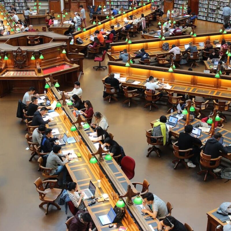 Biblioteca con tavoli lunghi e tanti studenti seduti mentre studiano e leggono.