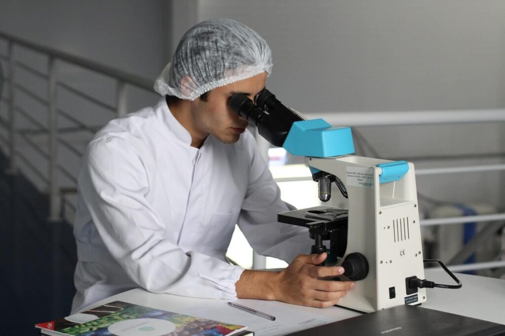 Biologo mentre guarda il microscopio in un laboratorio. Indossa un camice bianco e una cuffia per i capelli. Vicino a se ha una penna e un quaderno.