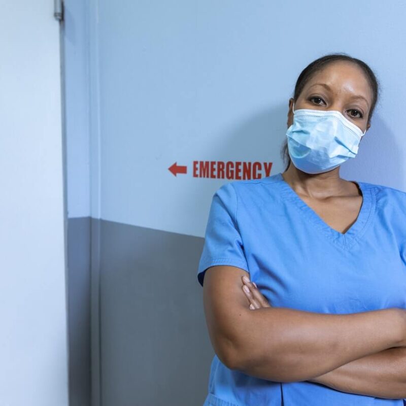 Medico con indosso uno scrub blu e una mascherina azzurra. È poggiata a un muro blu con dietro una scritta in rosso della parola "Emergency".