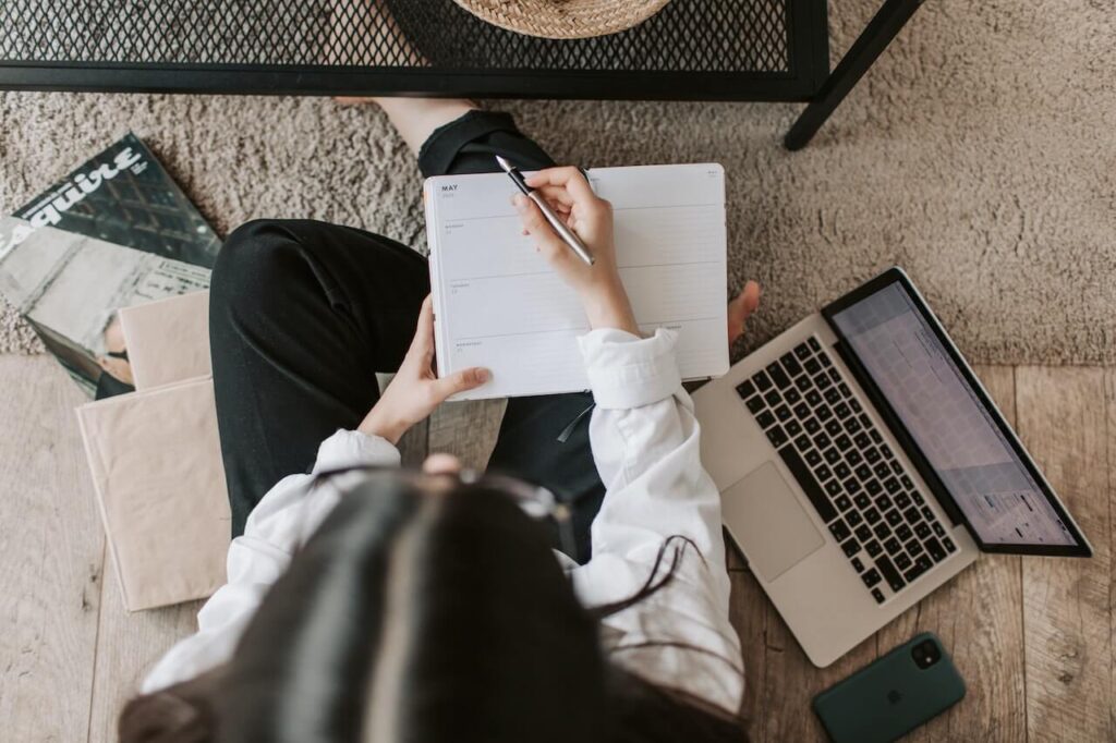 Ragazza seduta sul pavimento a gambe conserte mentre scrive sul diario e studia usando il pc.
