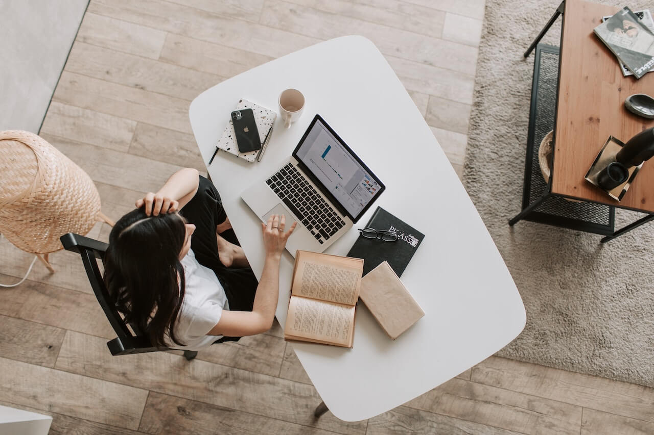Studentessa seduta davanti a una scrivania con sopra libri, pc e telefono.