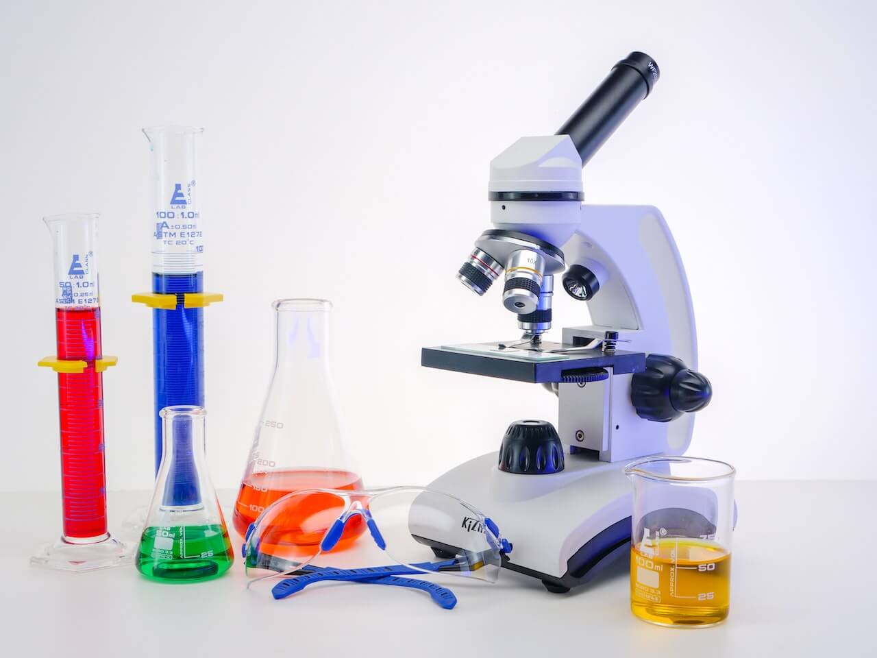 Microscopio circondato da provette e backer in un laboratorio bianco. Tra gli oggetti ci sono anche degli occhiali protettivi per gli esperimenti di laboratorio.