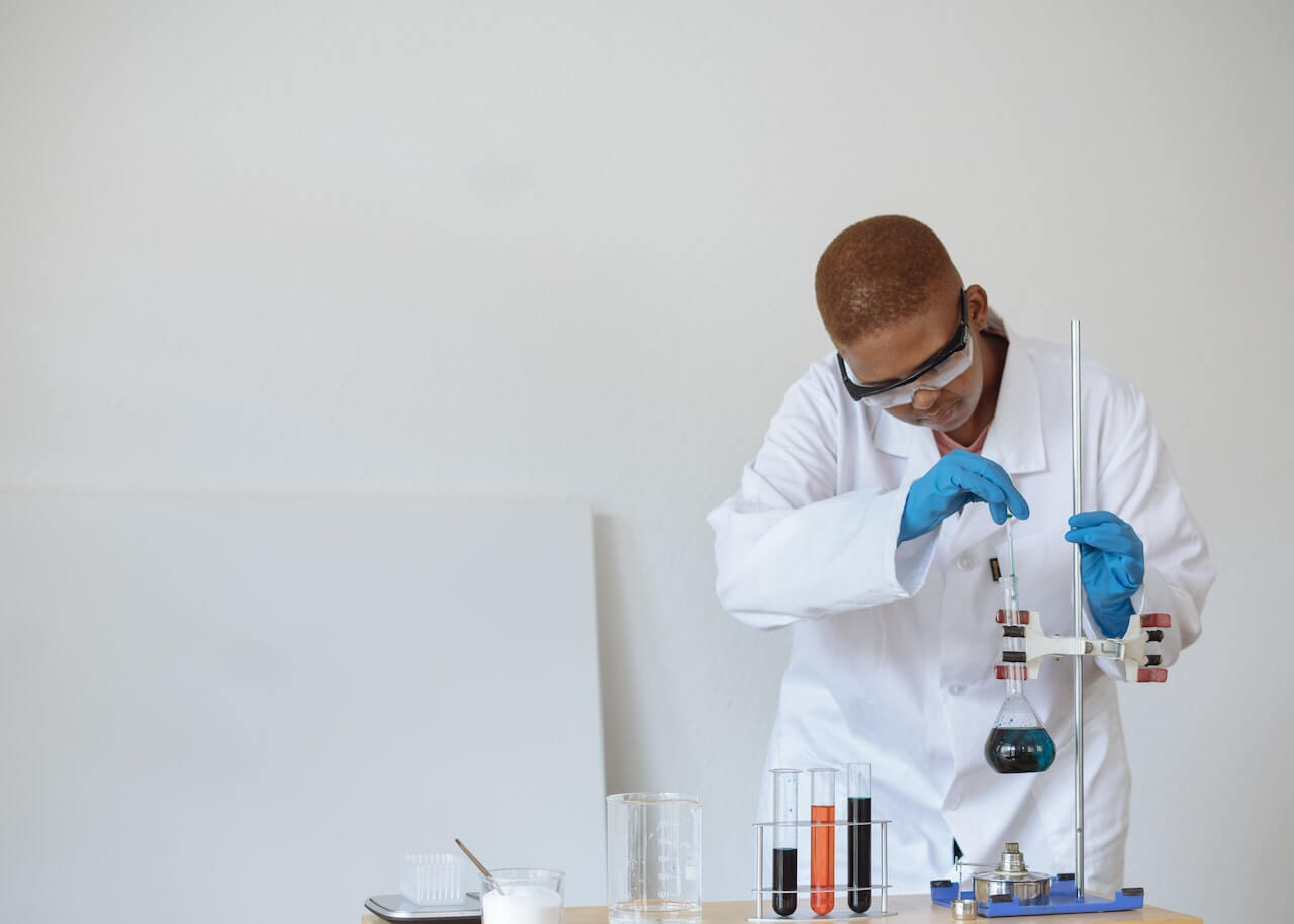 Biologo mentre effettua un esperimento in laboratorio con provette e beker. Indossa un camice bianco, occhiali protettivi e guanti blu.