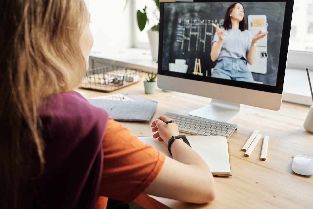 Studentessa seduta alla scrivania mentre prende appunti su un quaderno e guarda una lezione online dal suo pc.