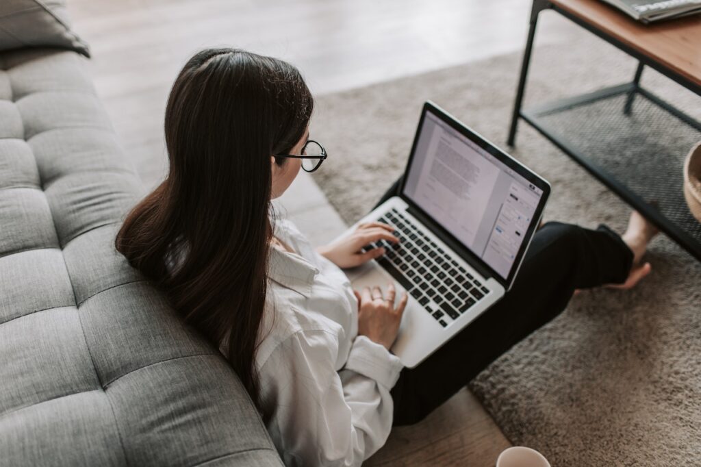 Ragazza mentre utilizza il pc poggiato sulle gambe. È distesa con le spalle appoggiate alla seduta del divano del salotto.