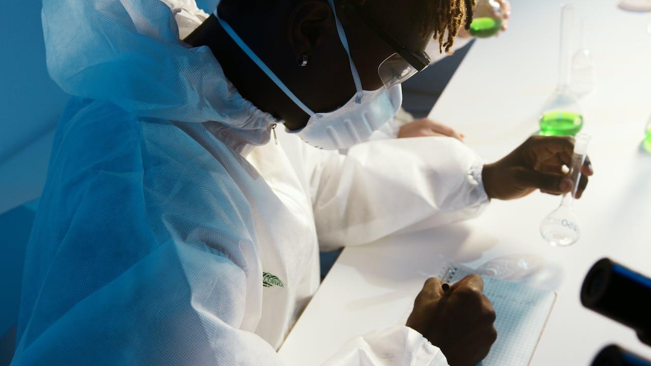Chimico in laboratorio che annota su un foglio le proprietà di un composto in un'ampolla che tiene nella mano sinistra.