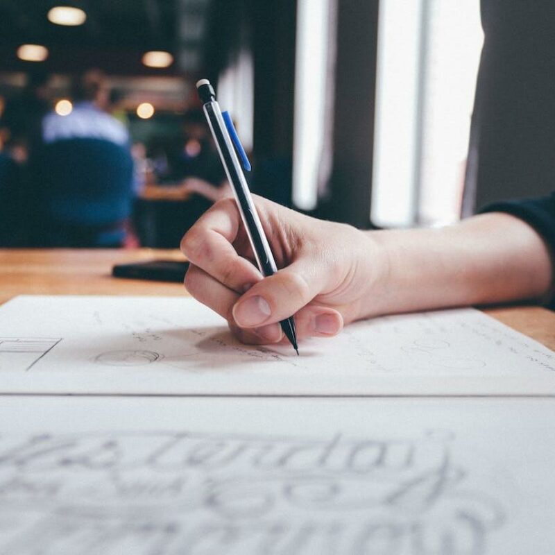 Mano che tiene in mano una penna mentre scrive su un foglio bianco poggiato su una scrivania.