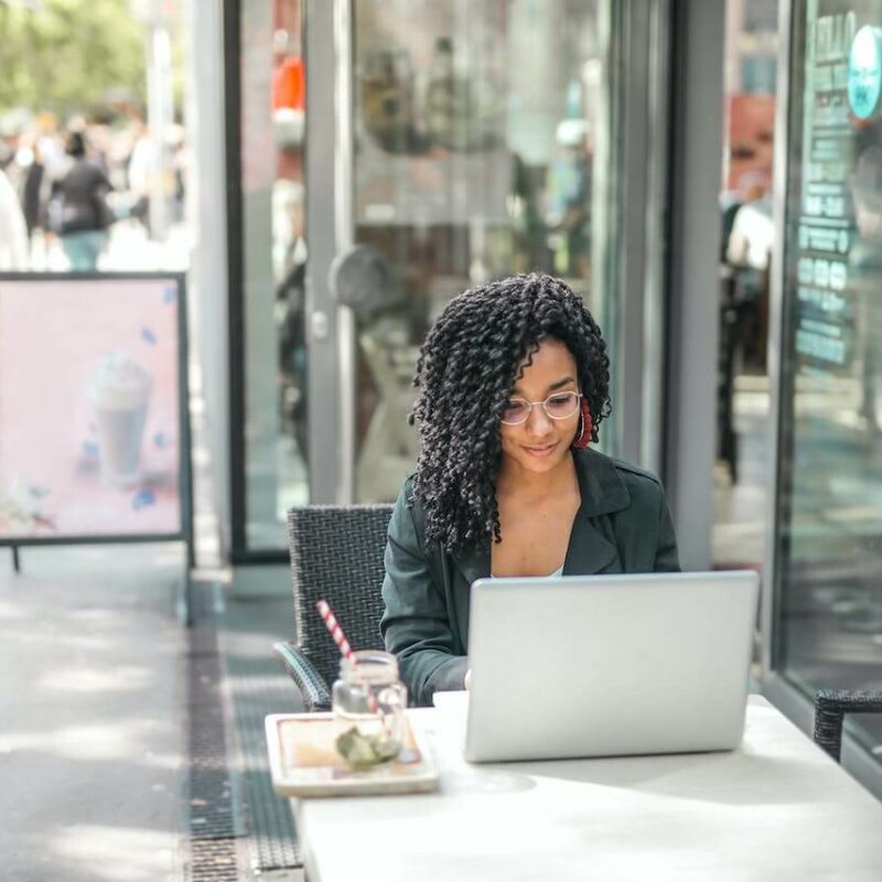 Ragazza seduta al tavolino esterno di un bar mentre lavora al computer e beve un drink.