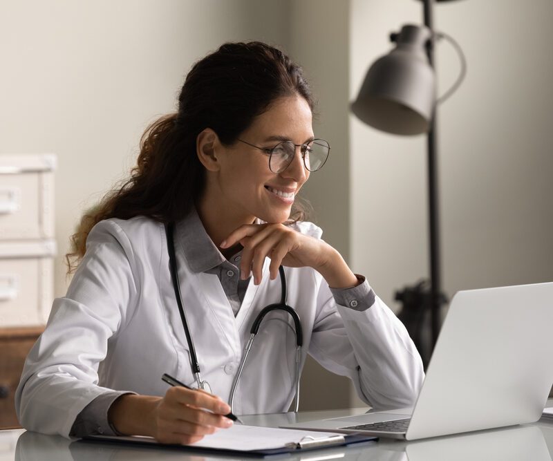 Un medico donna con occhiali e uniforme che riempe documenti sulla storia clinica del paziente, guarda lo schermo del laptop.