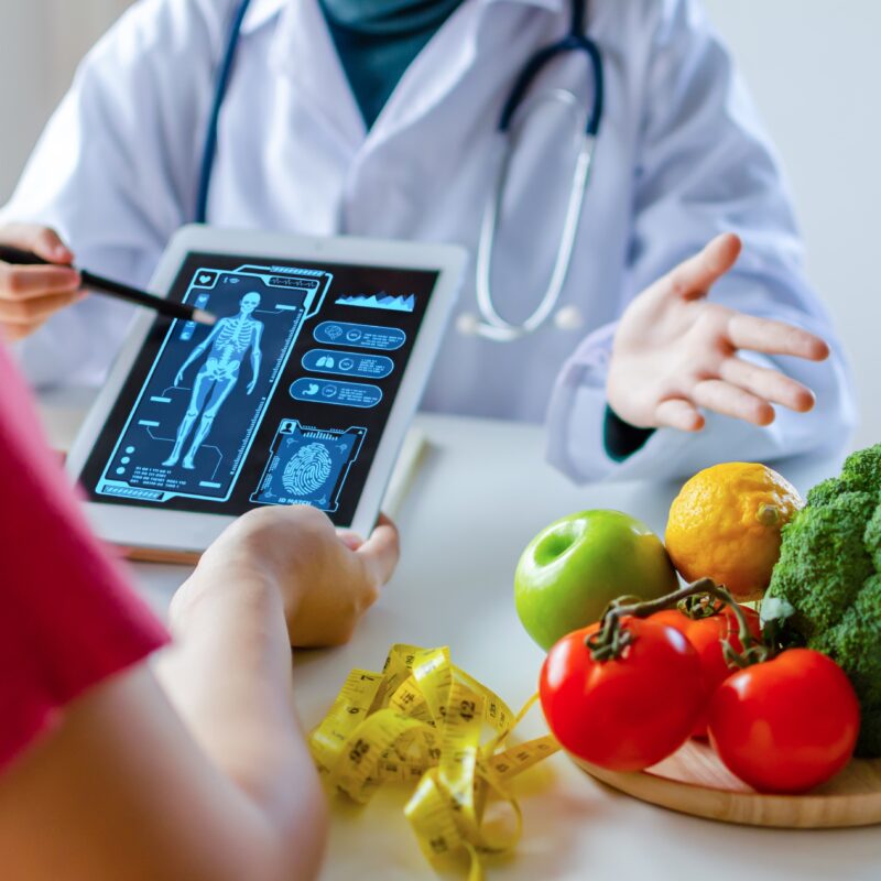 Medico specializzato in Scienza dell'Alimentazione che utilizza un tablet con diagramma di icone grafiche virtuali e verdura e frutta con paziente sulla scrivania in ospedale.