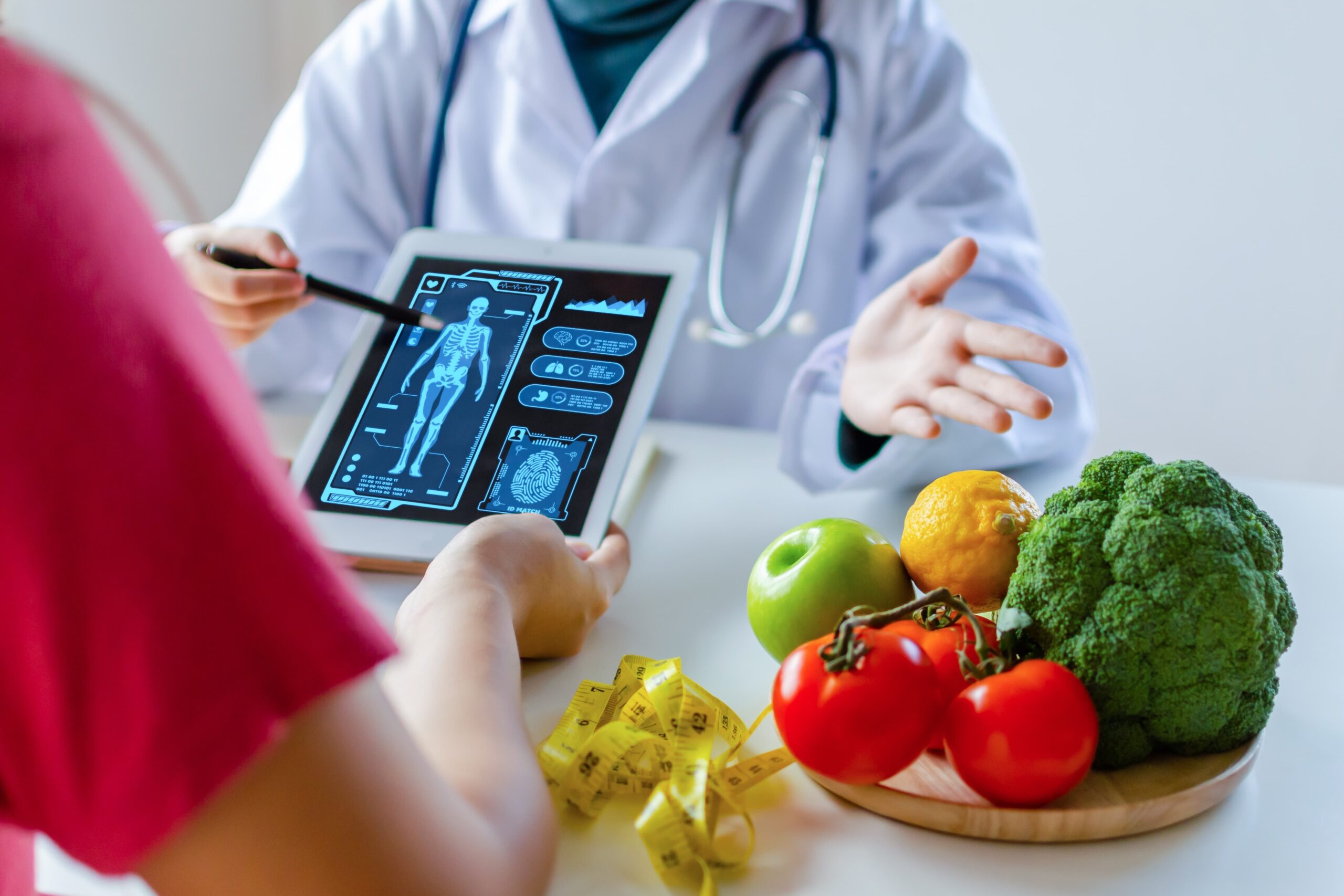 Medico specializzato in Scienza dell'Alimentazione che utilizza un tablet con diagramma di icone grafiche virtuali e verdura e frutta con paziente sulla scrivania in ospedale.