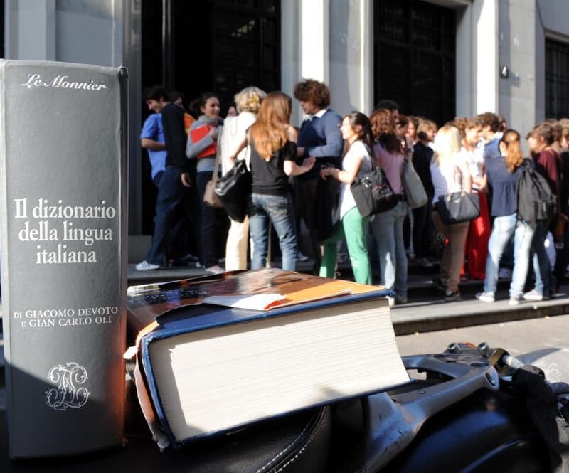 Studenti in attesa davanti a una scuola con dizionari in primo piano, simbolo dei collegamenti interdisciplinari per il colloquio orale della Maturità 2025