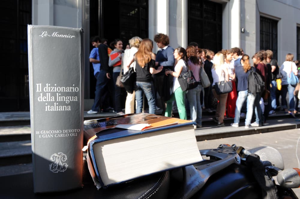 Studenti in attesa davanti a una scuola con dizionari in primo piano, simbolo dei collegamenti interdisciplinari per il colloquio orale della Maturità 2025