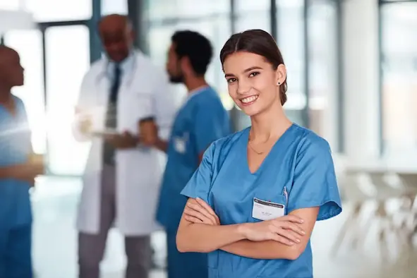 Studentessa in uniforme sanitaria sorride in primo piano, con colleghi e medici sullo sfondo in un ambiente ospedaliero moderno — rappresentazione della preparazione per il Test Professioni Sanitarie 2025.