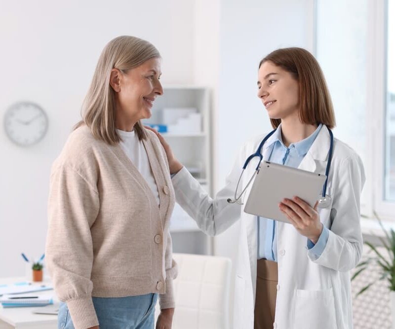 Una dottoressa sorridente dopo aver passato il concorso ssm 2025 in camice bianco e stetoscopio al collo tiene un tablet e ha una mano sulla spalla di una paziente, che sorride e la guarda.